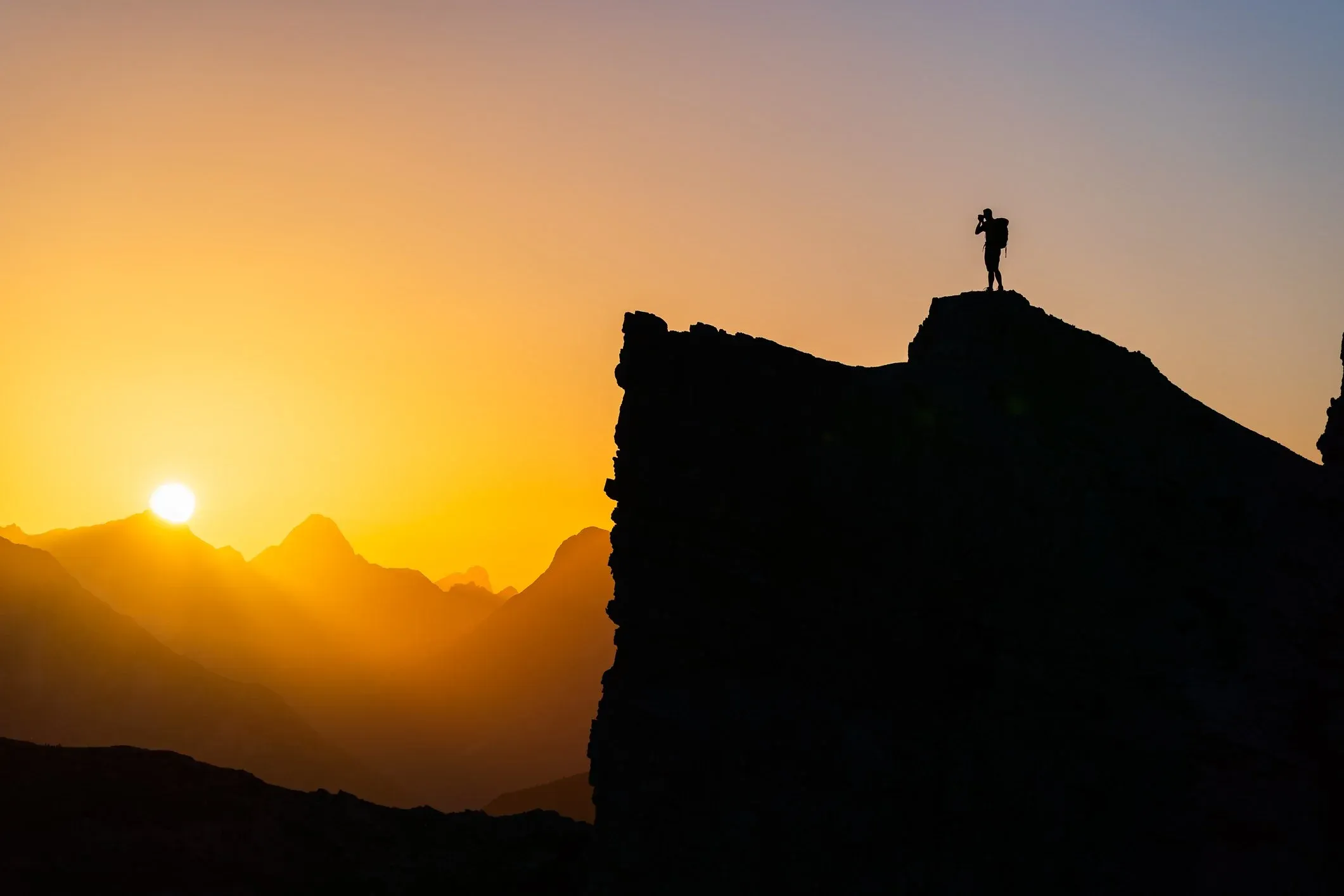 Silhouetted figure on a cliff edge as the sun sets over peaks.